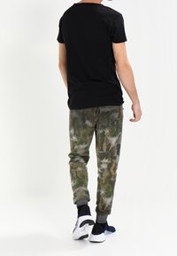 Schwarzes Kurzarm-T-Shirt, camouflierte Jogginghose in Grün und Beige, kombiniert mit marineblauen Sneakers. Rückansicht mit elastischen Bündchen an der Hose.
