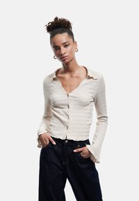 Lys beige struktureret bluse med krave, knaplukning og flagrende manchetter. Sat sammen med mørke denimbukser med høj talje.