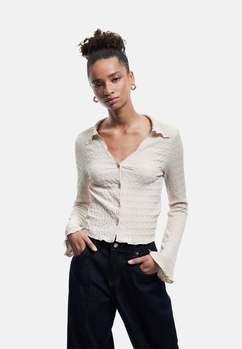 Lichtbeige textuurblouse met een kraag, knoopsluiting en uitlopende mouwen. Gecombineerd met een donkerblauwe hoge taillejeans.