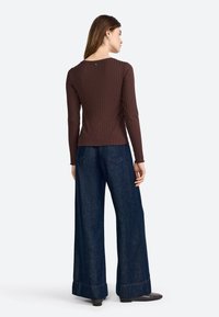 Top lungo a coste marrone con scollo rotondo e dettaglio dei bottoni sul retro, abbinato a jeans a gamba larga blu scuro con orli risvoltati.