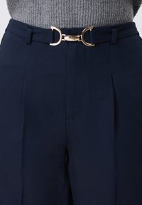 Pantalon taille haute bleu marine avec un design plissé, ceinturé d'une boucle circulaire dorée, accompagné d'un pull gris texturé.