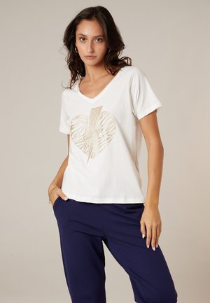 Woman wearing white V-neck shirt with gold heart and lightning bolt design and navy blue pants, standing with one hand in pocket.