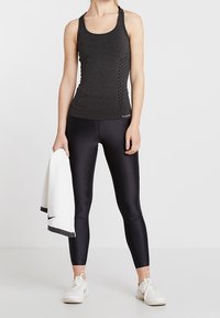 Canotta nera sportiva con pannello laterale testurizzato, abbinata a leggings neri. Asciugamano bianco con bordo nero tenuto in mano. Sneakers bianche.