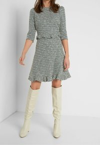 Vihreä ruudullinen mekko, jossa on kolme neljäsosapituista hihaa, vyötäröä korostava malli ja röyhelöhelma, yhdistettynä polveen ulottuviin valkoisiin nahkasaappaisiin.