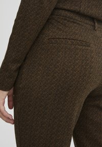 Brown herringbone patterned trousers in a textured fabric. Features a rear pocket, fitted shape, and visible stitching accents.