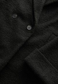 Schwarzer Wollmantel aus strukturiertem Stoff, mit zwei schwarzen Kunststoffknöpfen und klassischem Reversdesign. Nahaufnahme des Stoffes und der Details.