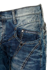 Blaue Denim-Jeans mit einem verblassten Finish, ausgestattet mit Kontrastnähten, mehreren Taschen und metallischen Nieten. Markenetikett sichtbar.