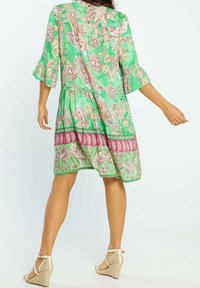 Vestido corto verde con patrones florales y paisley, mangas con volantes y falda en capas; acentuado con un borde rosa y verde en el dobladillo.