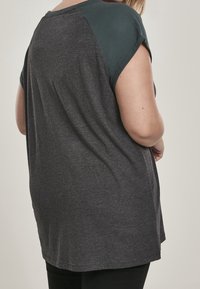 Camiseta gris con un panel de hombro verde oscuro, de manga corta, corte relajado y textura suave; costuras visibles en la parte posterior.