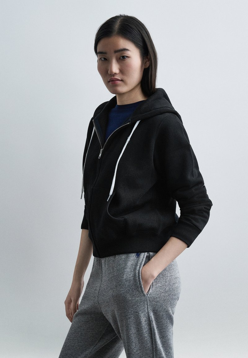 Νεαρή γυναίκα που φοράει μαύρη φούτερ ζακέτα με  φερμουάρ πάνω από μπλε πουκάμισο και ανοιχτό γκρι φόρμα, στέκεται με το ένα χέρι στην τσέπη μπροστά σε απλό φόντο.