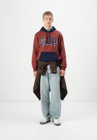 Jeune homme portant un hoodie GAP rouille et marine, un jean large bleu clair, des baskets blanches et une veste noire nouée autour de la taille.