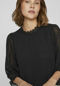 Sort bluse med struktureret polka dot-mønster, blondertrimmet halsudskæring og gennemsigtige pufærmer. Blødt stof med en glat finish.
