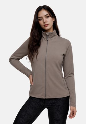 Young woman with long dark hair wearing a taupe zip-up jacket and black patterned leggings, posing with one hand on her hip.