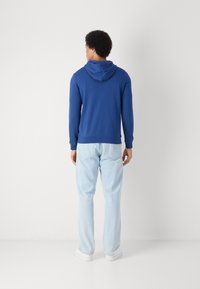 Felpa blu con cappuccio, cordino e tasca a marsupio, abbinata a jeans chiari. Design semplice, vestibilità casual e texture morbida.