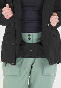 Mustapuku, jossa on vetoketju ja napitus, päällä vaaleanvihreät housut, joissa on nappi ja joustava vyötärö.