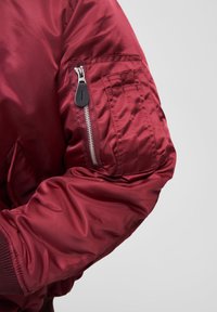 Veste bomber bordeaux avec finition brillante, dotée d'une poche zippée sur la manche, de poignets côtelés et de détails en tissu texturé.