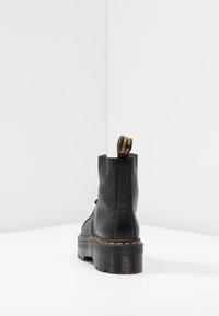 Bottines en cuir noir avec une semelle épaisse crantée et des coutures jaunes, vues de l'arrière sur une surface blanche.
