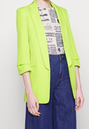 Persoon die een limoengroene blazer draagt over een top met krantenprint en hoge taille, donkerblauwe wijde broekspijpen, staand tegen een witte achtergrond.