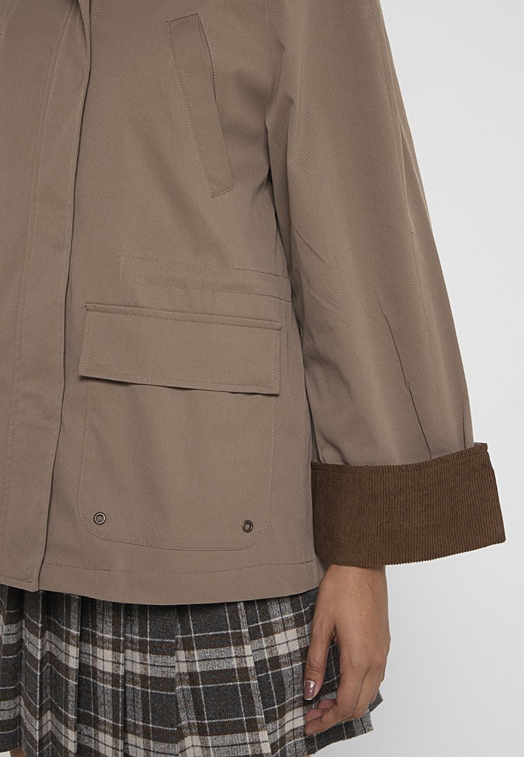  lichtbruine jas met een getextureerde corduroy manchette, voorzien van twee zijzakken en een rechte snit. De stof lijkt glad en duurzaam.