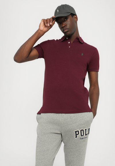 Bordo polo majica s zelenim logotipom, kratkim rukavima i ovratnikom, u kombinaciji sa sivim trenerkama s natpisom "POLO".