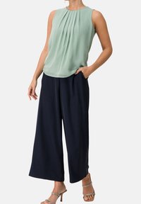 Blusa sin mangas de color verde claro con frente plisado, combinada con pantalones anchos azul marino y sandalias de tacón con tiras plateadas.