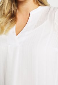 Donna bionda che indossa una blusa bianca a V con una sottile texture a strisce verticali, capelli che cadono su una spalla, primo piano su collo e petto.