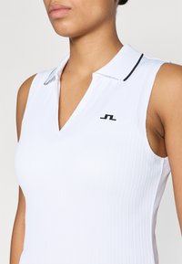 Bezruký bílý polo top s žebrovanou texturou, se černým límečkem a malým černým logem na hrudi.