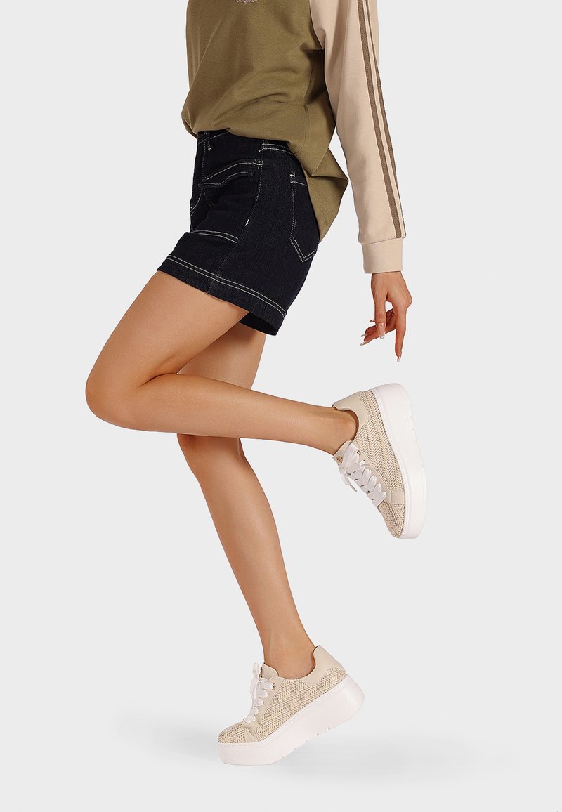 Sneaker con una toma in beige testurizzato e suola piatta bianca, abbinate a pantaloni corti in denim scuro. L'outfit presenta una maglietta verde oliva e beige.