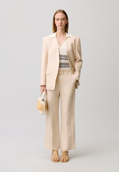 Femme en complet pantalon beige, gilet rayé, tenant un petit sac à main tressé, portant des sandales jaunes à lacets, debout devant un fond gris.