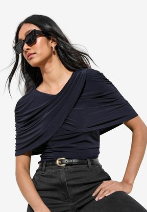 Frau trägt schwarzen gerafften schulterfreien Oberteil, schwarzen Gürtel mit goldener Schnalle, schwarze Hose, goldene Creolen und übergroße schwarze Sonnenbrille.