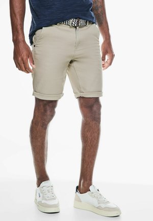 Beige katoenen shorts met opgerolde mouwen, voorzakken en een zwart-wit geblokte riem. Gecombineerd met witte sneakers met grijze accenten.