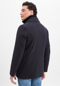 Mann in schwarzem Wollmantel und blauen Jeans, seitlich gedreht, der leicht zurückschaut, vor schlichtem weißen Hintergrund.