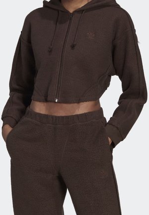 Henkilö, joka pukeutunut tummanruskeaan lyhennettyyn vetoketjulliseen huppariin ja siihen sopiviin, kuvioituihin verkkarihousuihin, kädet taskuissa, yksivärisellä taustalla.