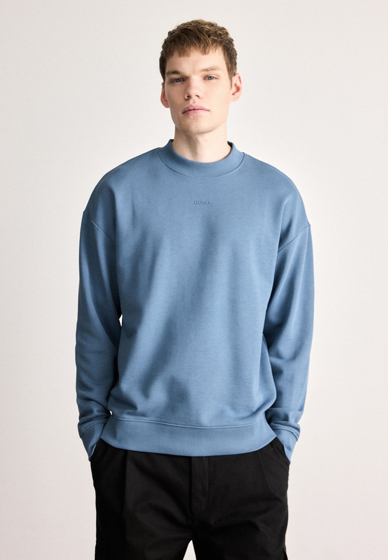 Ανοιχτό μπλε φούτερ με ριμπ λαιμόκοψη, φαρδιά εφαρμογή και διακριτική λεπτομέρεια λογότυπου στο μπροστινό μέρος, συνδυασμένο με μαύρο παντελόνι.