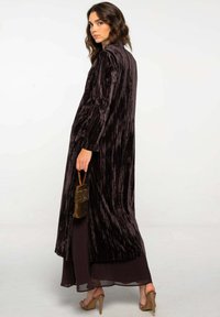 Manteau long en velours violet foncé avec des rayures verticales, manches longues, fentes sur les côtés, superposé sur une robe sombre transparente, associé à un sac à main scintillant.