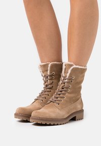 Botas de tobillo de ante color marrón claro con un suave forro de piel sintética blanca en la parte superior, diseño tradicional de cordones y suelas de goma marrón resistente.