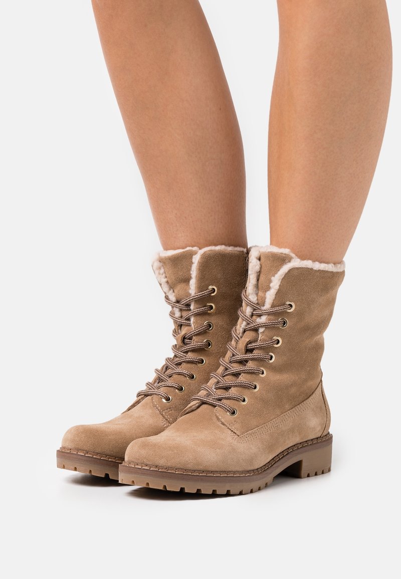 Botas de tobillo de ante color marrón claro con un suave forro de piel sintética blanca en la parte superior, diseño tradicional de cordones y suelas de goma marrón resistente.