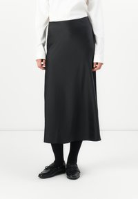 Schwarzer Midirock aus glattem Stoff, mit einem schlichten Design, gerader Silhouette und ohne sichtbare Muster. Kombiniert mit schwarzen Schuhen und Socken.