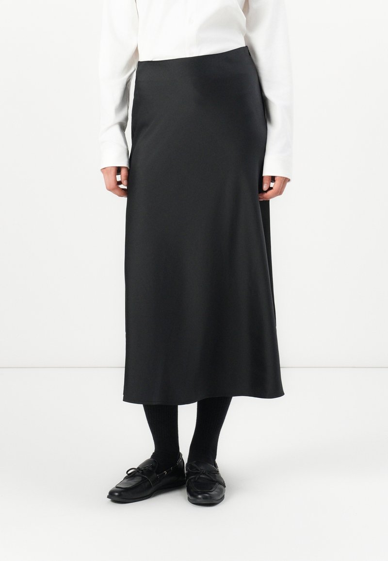 Schwarzer Midirock aus glattem Stoff, mit einem schlichten Design, gerader Silhouette und ohne sichtbare Muster. Kombiniert mit schwarzen Schuhen und Socken.