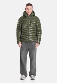 Zielona pikowana kurtka z kapturem, zapinana na zamek z przodu, z poziomym pikowaniem. W zestawie z szarymi spodniami dżinsowymi i białymi trampkami.