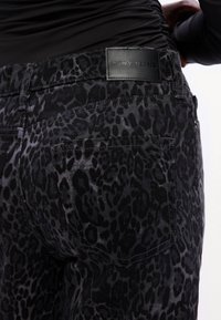 Melna denima bikses ar leopardu rakstu, ar ādas jostas uztveri, uz kuras ir uzraksts "DKNY Jeans", un aizmugurējās kabatas detaļa.