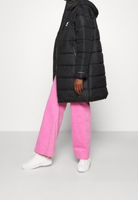 Abrigo puffero negro con capucha y costuras horizontales, combinado con pantalones de pierna ancha rosas y zapatillas blancas, presentado sobre un fondo neutro.