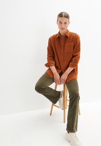 Chemise en velours côtelé orange surdimensionnée, poche avant, manches retroussées, associée à un pantalon évasé vert olive et des baskets blanches sur un tabouret en bois.