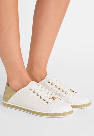 Fødder iført hvide lærredssneakers med guldfarvede øjer og detaljer, fletdet jute kant langs sålen, på en ensartet lys baggrund.