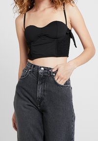Černý zkrácený top s lodičkovým výstřihem, nastavovacími ramínky a zdrsněnými bočními panely. V kombinaci s šedými džíny s vysokým pasem.