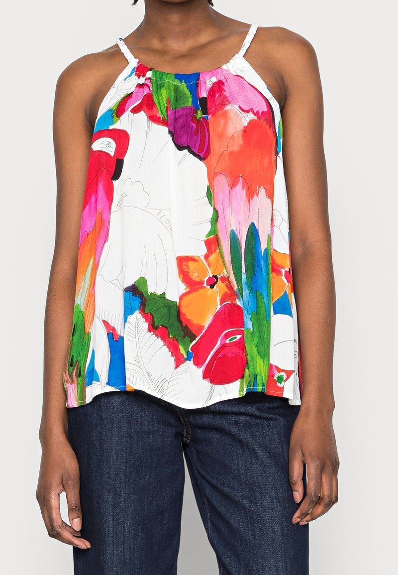 Femme portant un haut blanc sans manches avec des imprimés floraux et d'oiseaux colorés, associé à un jean bleu foncé.