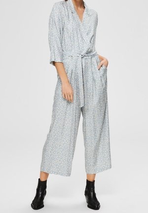 Lichtblauwe bloemenjumpsuit met een wikkelontwerp, driekwartmouwen, een ceintuur in de taille, wijde pijpen en zijzakken. Gepaard met zwarte enkellaarsjes.