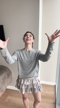 Joven mujer con suéter gris y falda beige con volantes está de pie en el interior con los brazos levantados, mirando hacia arriba con una expresión facial expresiva y esperanzada.