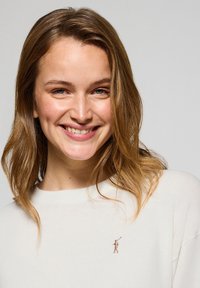 Gräddfärgad tröja med rund hals och ribbade mudd. Tröjan har en liten broderad logotyp på vänster bröst. Mjuk textur.