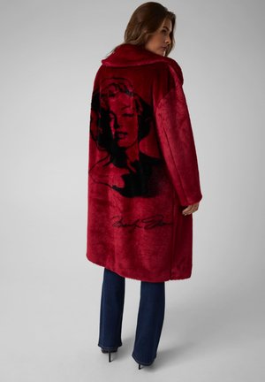 Une femme regarde par-dessus son épaule. Elle porte un long manteau rouge orné d’un grand portrait noir imprimé, un jean foncé et des chaussures à talons, sur un fond gris uni.
